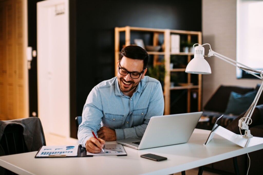 Homem escrevendo em papel impresso em seu home office; ao lado, luminária, tablet, notebook e celular