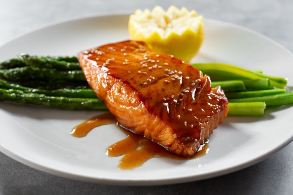 Em prato branco, salmão grelhado com molho teriyaki, aspargos e limão para decorar