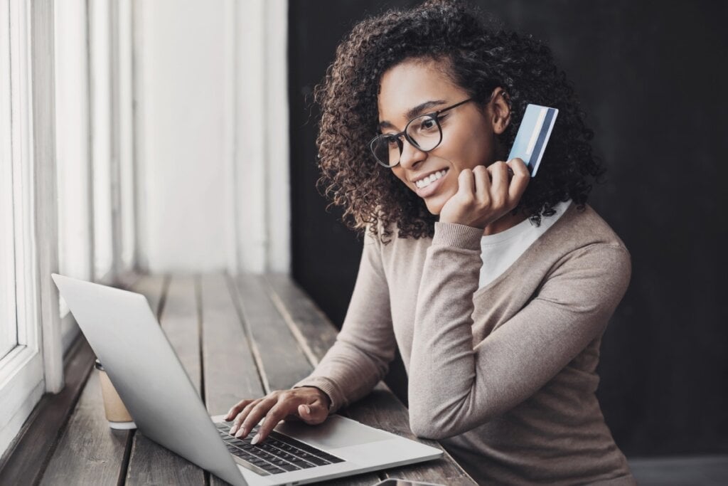 Mulher usando óculos sentada segurando um cartão e mexendo em um notebook