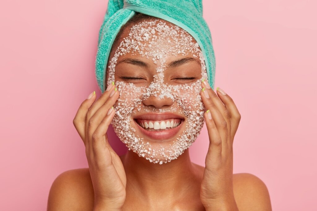 Mulher usando toalha verde na cabeça e sorrindo com as mãos no rosto fazendo esfoliação