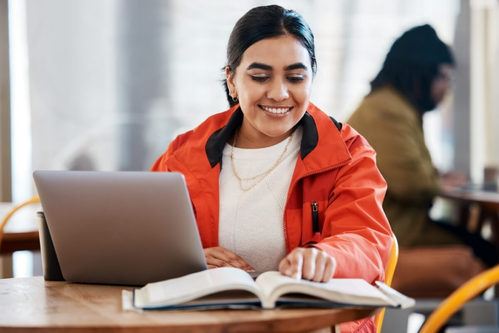 menina sorrindo sentada usando notebook e lendo livro ao mesmo tempo