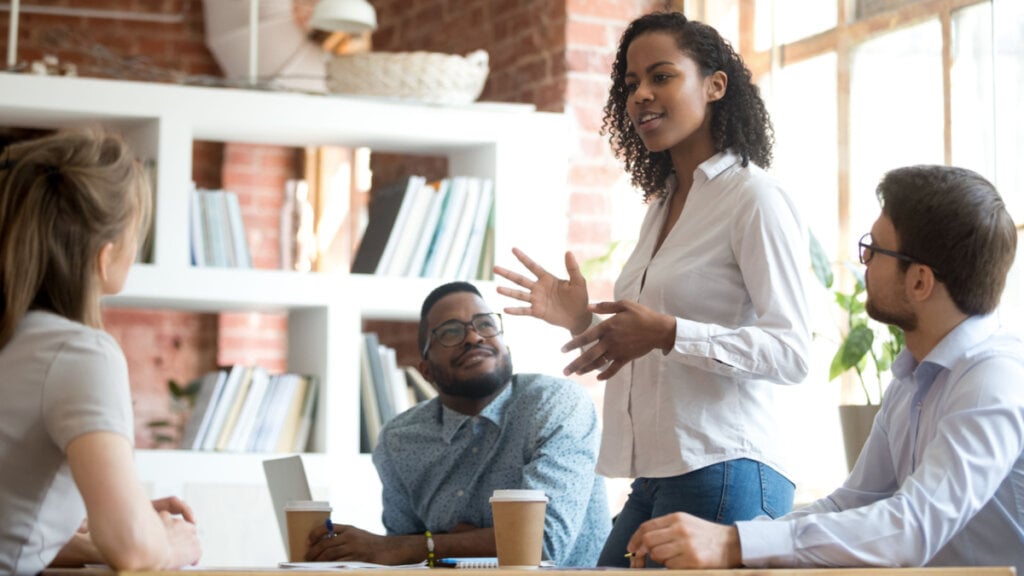 Mulher em pé se comunicando com outros colegas de trabalho, que estão sentados