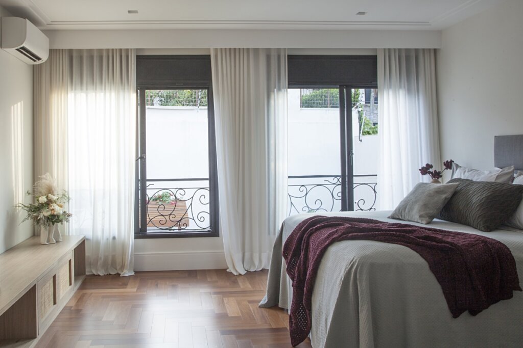 Quarto moderno e aconchegante com piso de madeira, grandes janelas com cortinas leves em tons neutros, cama com roupa de cama elegante e decoração minimalista