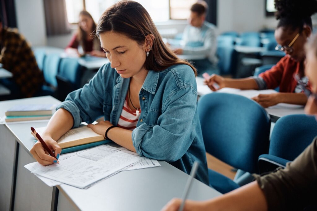 estudante em sala de aula fazendo uma prova, ela está com uma camisa jeans e uma camiseta listrada branca e vermelha