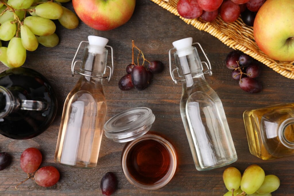 Três recipientes de vidro com vinagre de tipos diferentes em cima de uma mesa de madeira com frutas ao redor. 