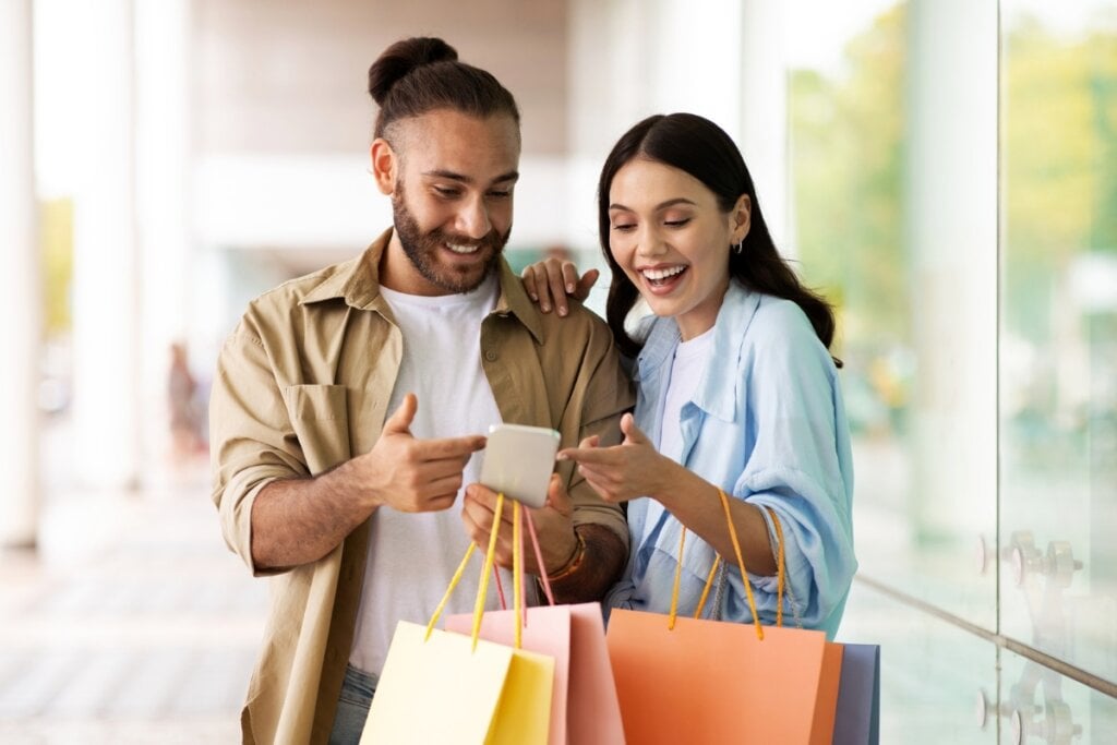 Homem e mulher um ao lado do outro segurando sacolas de compras, sorrindo e mexendo em um celular