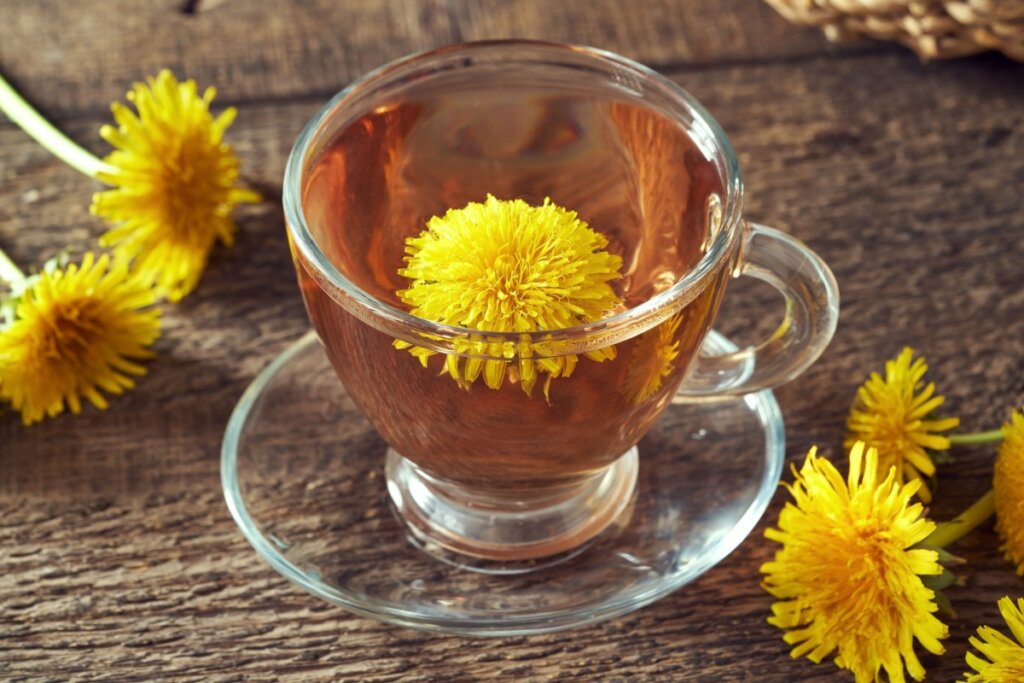 Chá de dente-de-leão servido em xícara de vidro com uma flor amarela dentro da xícara e em cima de uma mesa de madeira