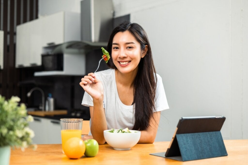 Mulher sorrindo, apoiada em mesa de madeira e segurando garfo com salada. o pote de salada branco está em cima da mesa