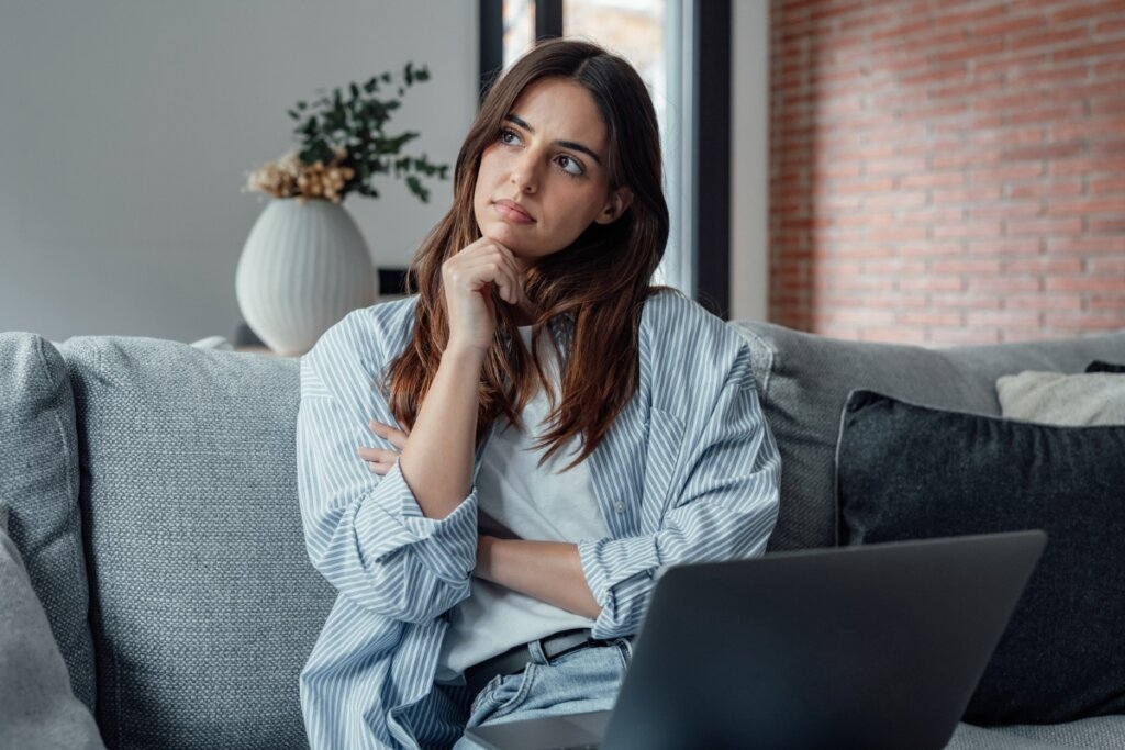 Mulher sentada em um sofá com um notebook no colo e a mão no queixo pensando 