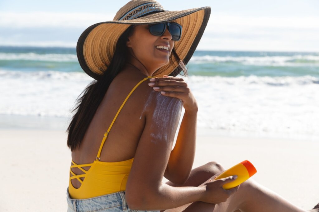 Mulher na praia passando protetor solar no braço, usando óculos escuro e chapéu 